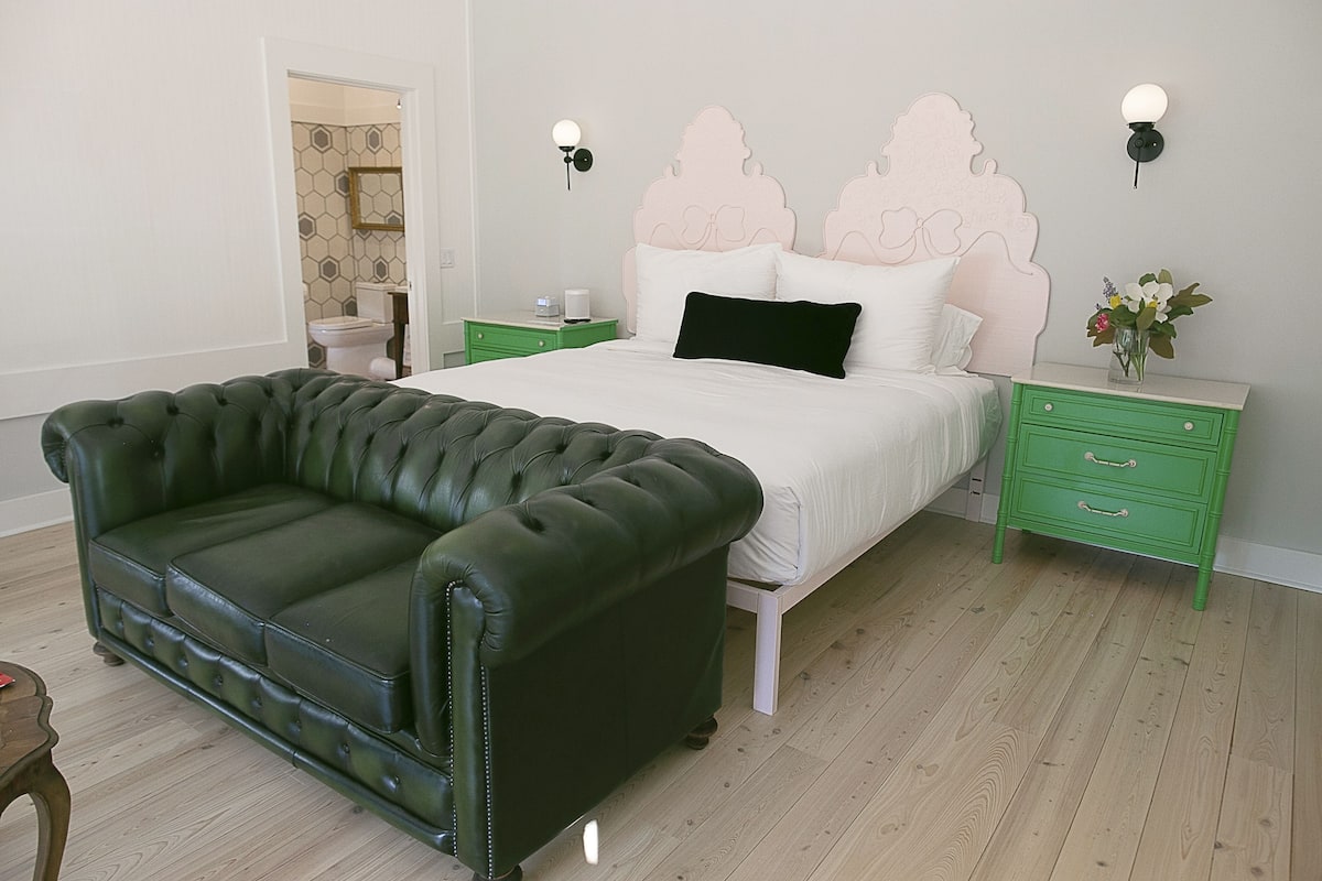 A bedroom with a green leather sofa, a bed with a pink headboard, green nightstands, and a wooden floor.