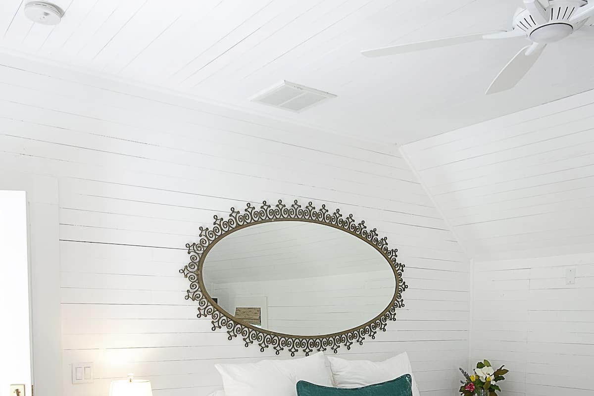 A bedroom with a white bed, a wooden nightstand, a mirror with an ornate frame, and a ceiling fan.