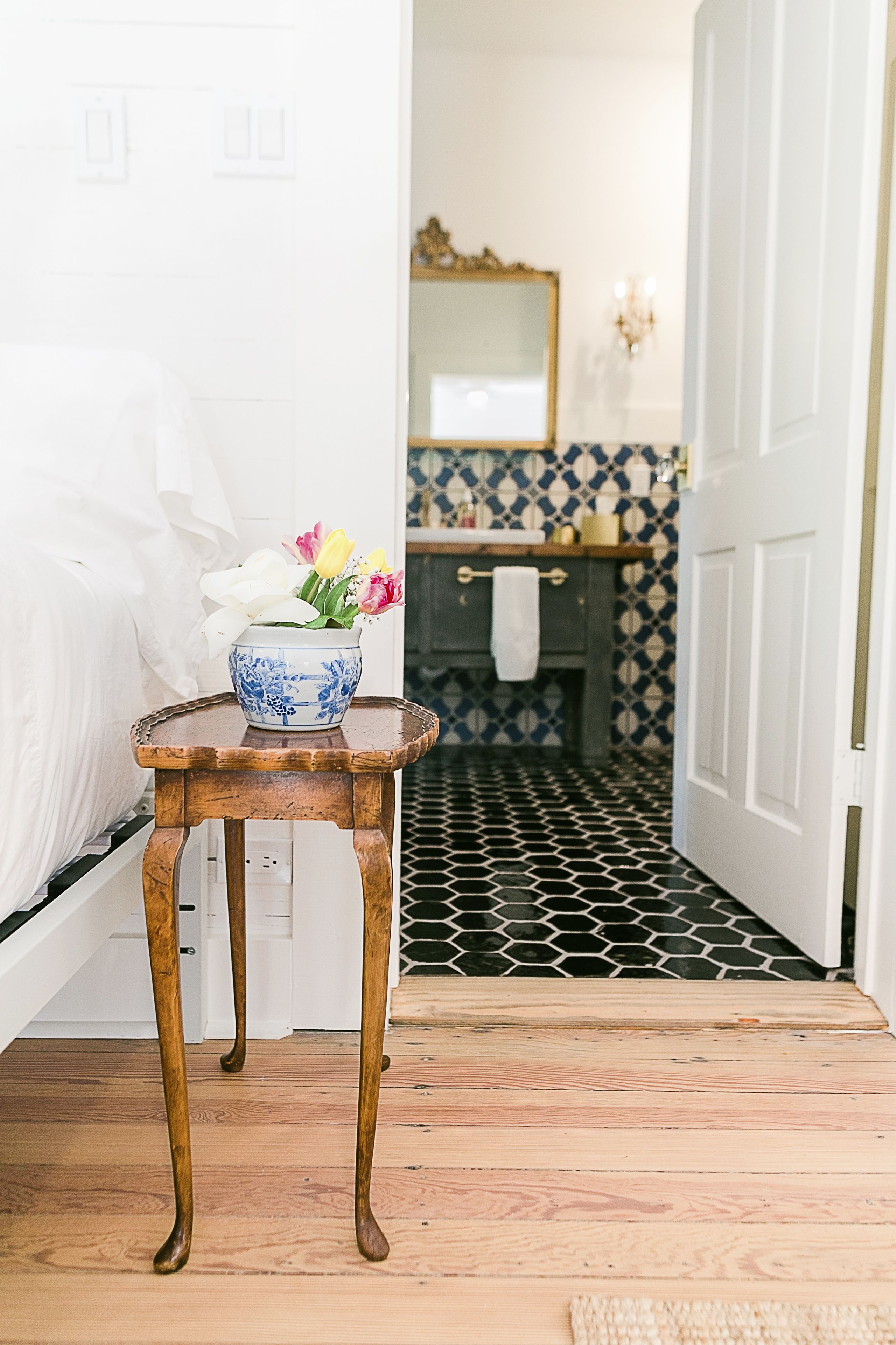 Un tabouret en bois avec un vase de fleurs, dans une salle de bain dont le sol est carrelé et la porte est blanche.