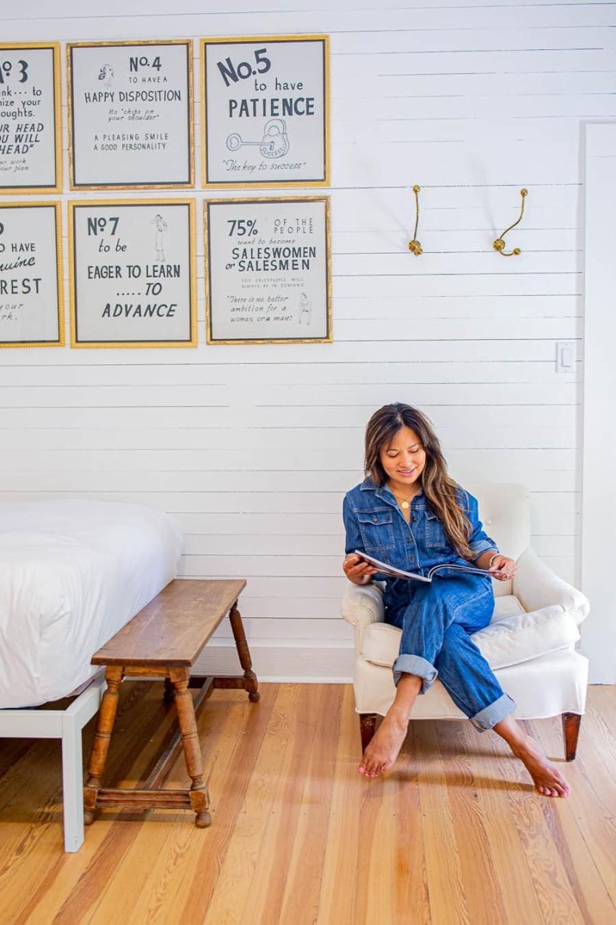 A person sitting on a white chair in a room with wooden flooring, a bed with white bedding, and motivational posters on the wall.