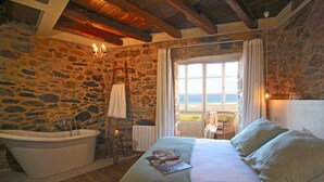 A bedroom with a stone wall, a bathtub, a bed with pillows, a window with a view, and a chandelier. - Unique oceanfront retreat with fantastic views of the wild natural space (Carballo)
