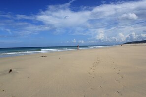Uma extensa praia de areia com pegadas que se estendem em direção à água, uma pessoa ao longe e céu claro com nuvens.