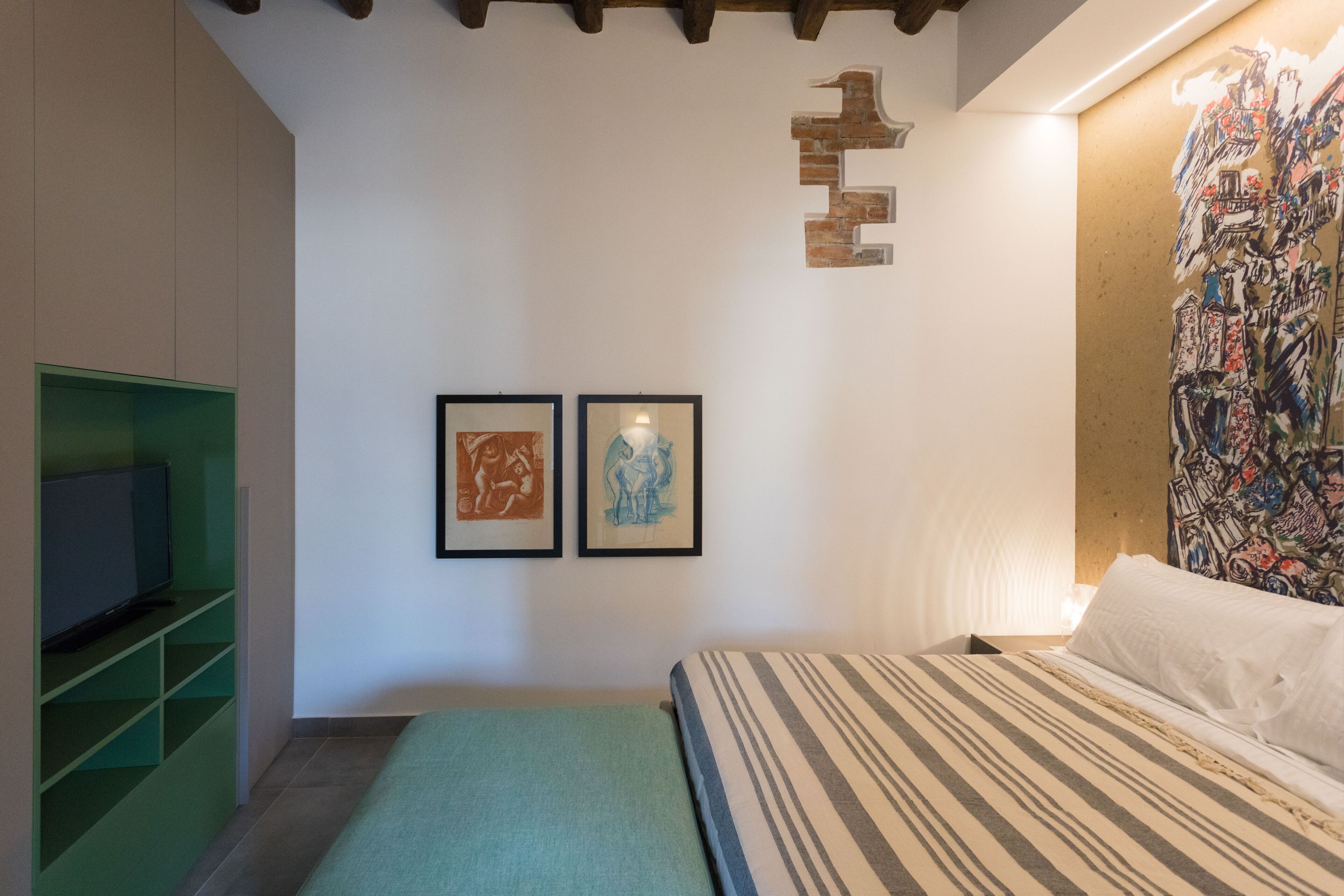 A bedroom with a bed, a green shelving unit, a television, and two framed artworks on the wall.