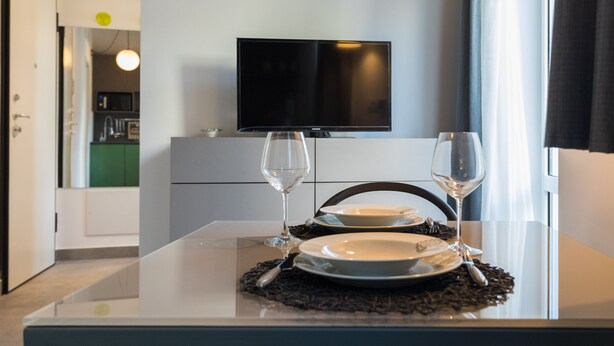 A dining table with two wine glasses and plates, a TV mounted on the wall, and a view of a kitchen area. - Suite Sant 'Agostino behind the cathedral (Palermo)