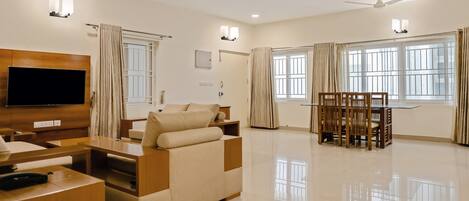 Una sala de estar moderna con un televisor de pantalla plana, un sofá, una mesa de comedor con sillas y ventanas grandes con cortinas.