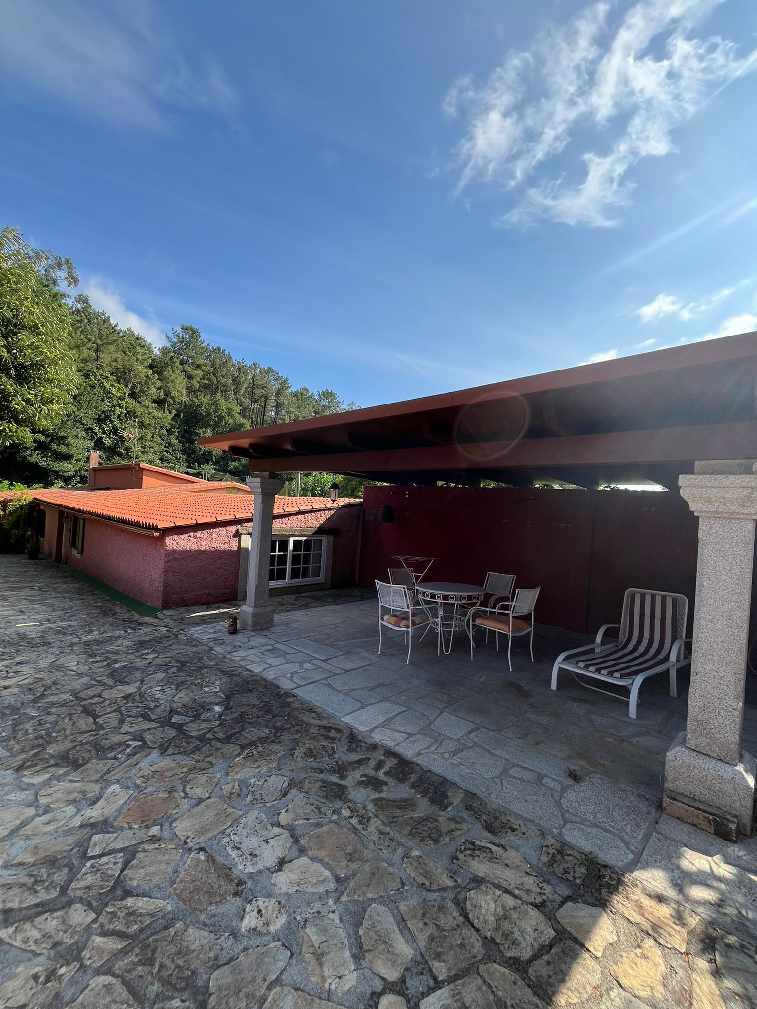 Un patio de piedra con mobiliario exterior, un edificio de techo rojo y un área cubierta con una columna de piedra.
