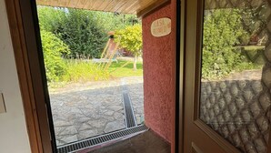 A wooden door leading to a stone-paved area with a metal grate, a red wall with a plaque, and a view of greenery outside. - CASA ROMEO housing in typical Galician architecture (Brión)