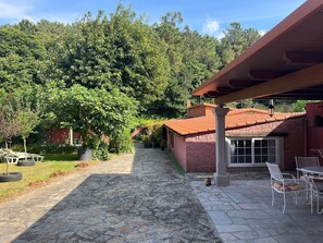 A stone-paved patio with a covered seating area, a house with a red-tiled roof, and a lush green forest in the background. - CASA ROMEO housing in typical Galician architecture (Brión)