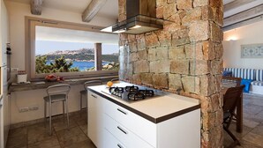 A kitchen with a stone wall, a gas stove, and a window overlooking a scenic view. - Charming villa with magnificent sea view (Santa Teresa di Gallura)