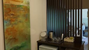 Una kitchenette compacta con refrigerador, un lavamanos pequeño y un espejo. Un cuadro abstracto de gran tamaño adorna la pared, y hay un mueble oscuro con toallas dobladas.