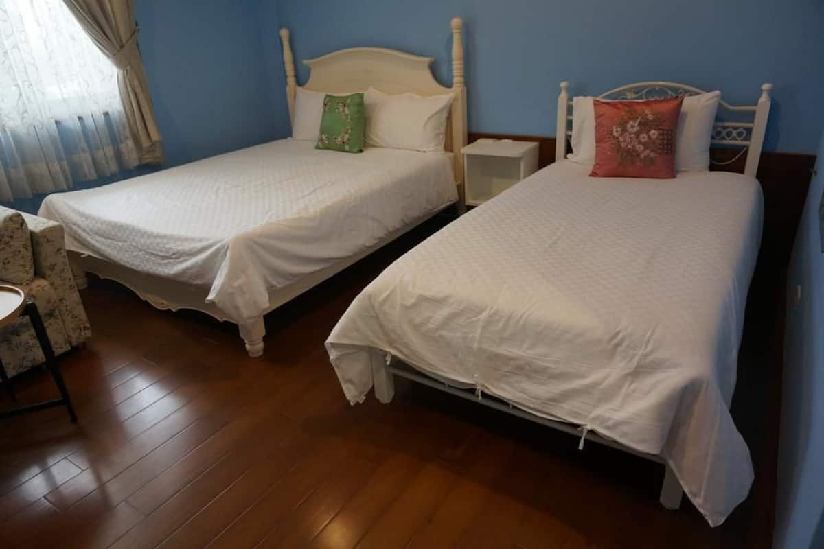 Deux lits simples avec du linge de lit blanc, une table de chevet en bois et une chaise à motifs floraux dans une pièce au plancher en bois et aux murs bleus.