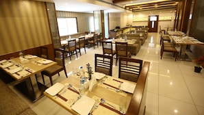 A hotel dining area with multiple tables set for dining, equipped with chairs, and a bar area in the background. - West Fort Hotel (Bengaluru)