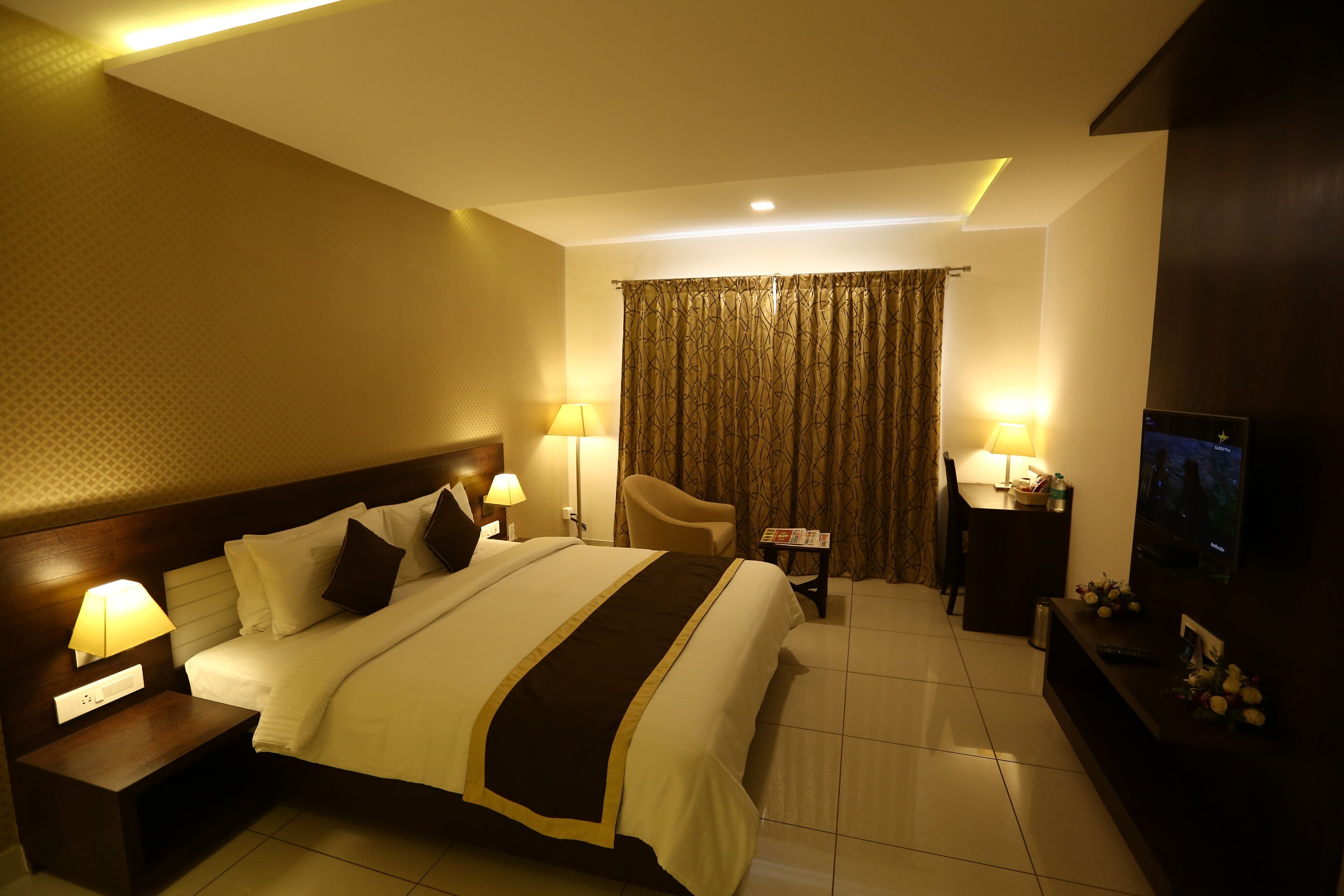 A hotel room with a large bed, two bedside lamps, a TV, a small table with a lamp, and a chair.