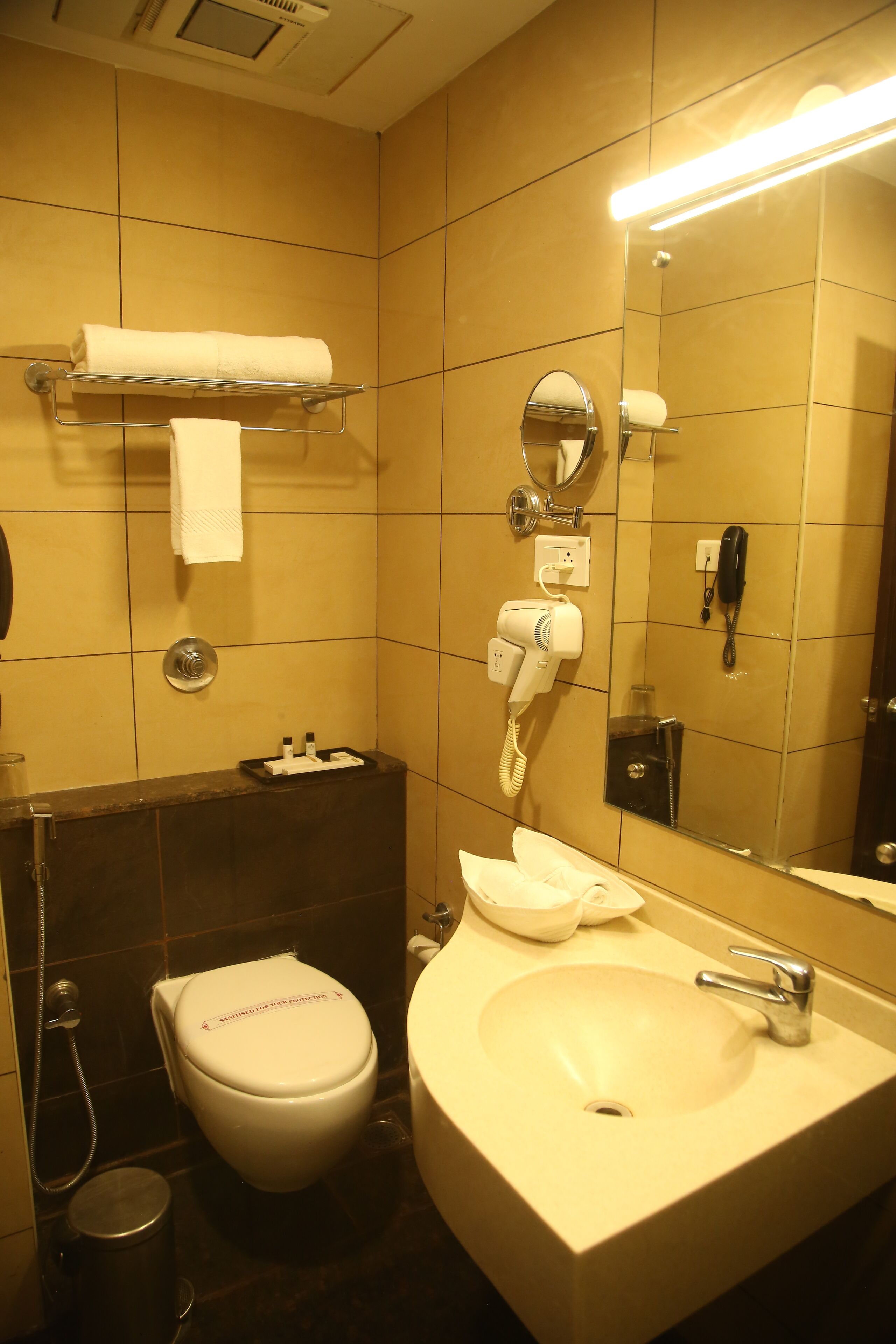 A bathroom with a toilet, sink, and mirror. There is a towel rack with a towel, a hairdryer, and a phone on the wall.