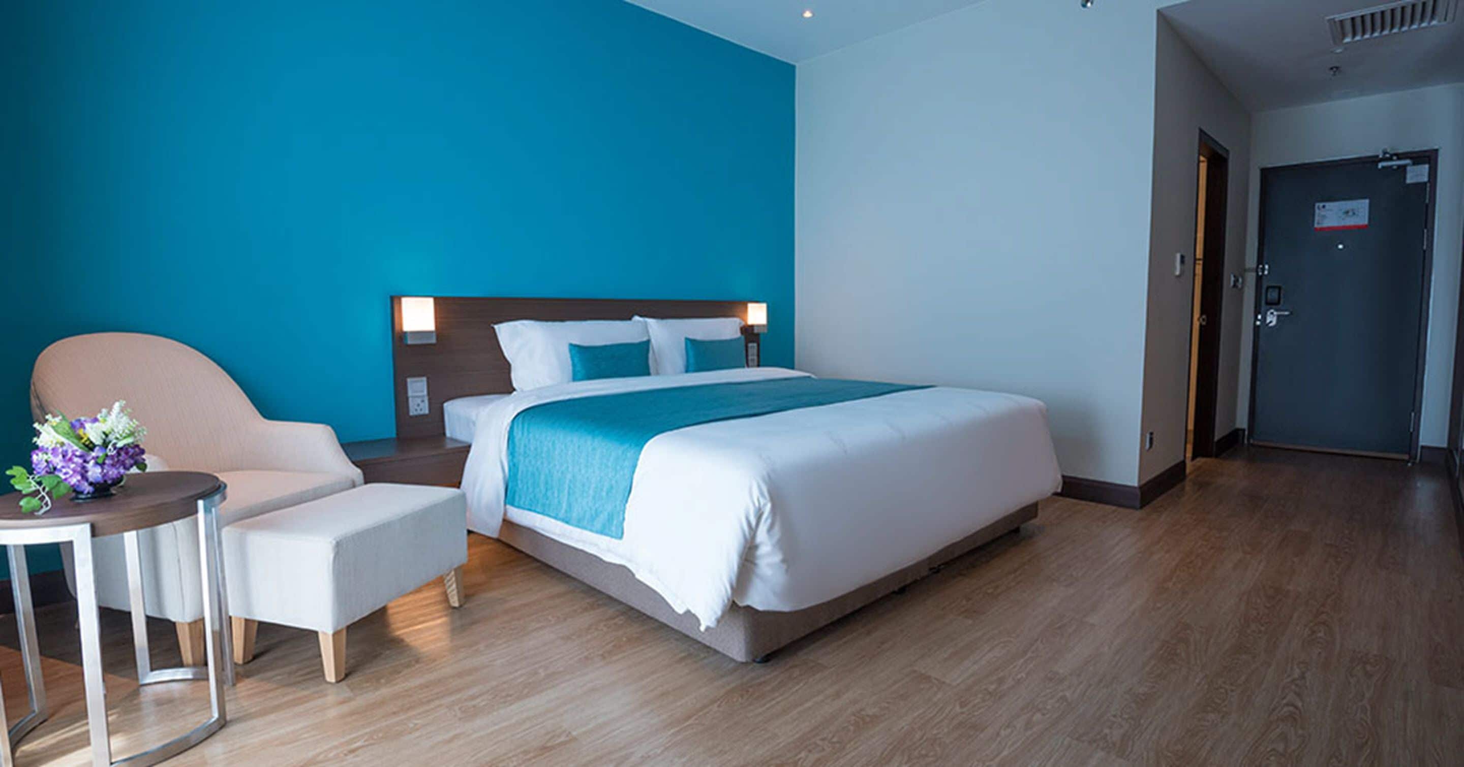 A modern hotel room with a large bed, a bedside table with a vase of flowers, a seating area with a chair, and a door to the right.