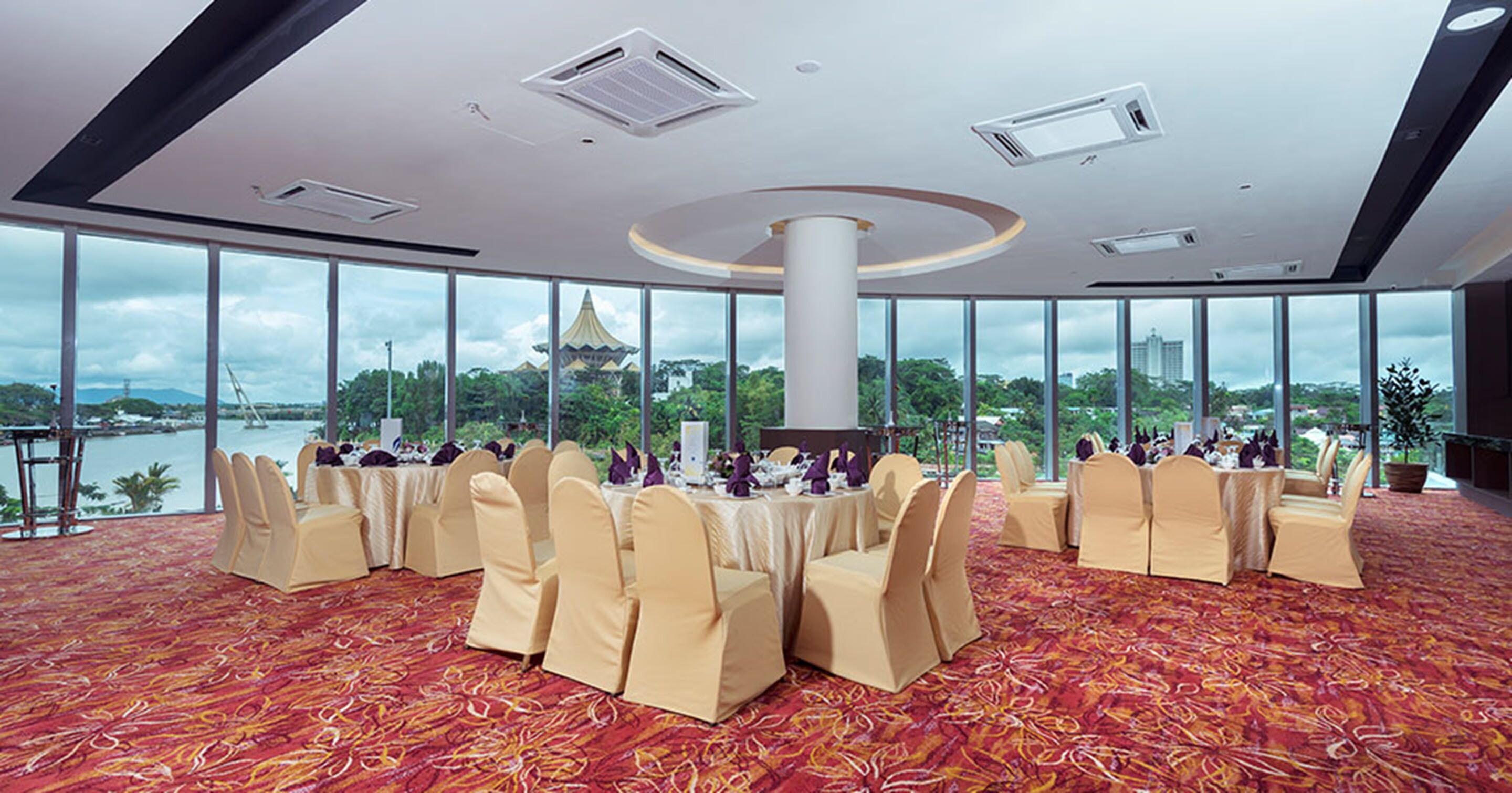 A banquet hall with round tables, beige chairs, and a view of a temple and water body.