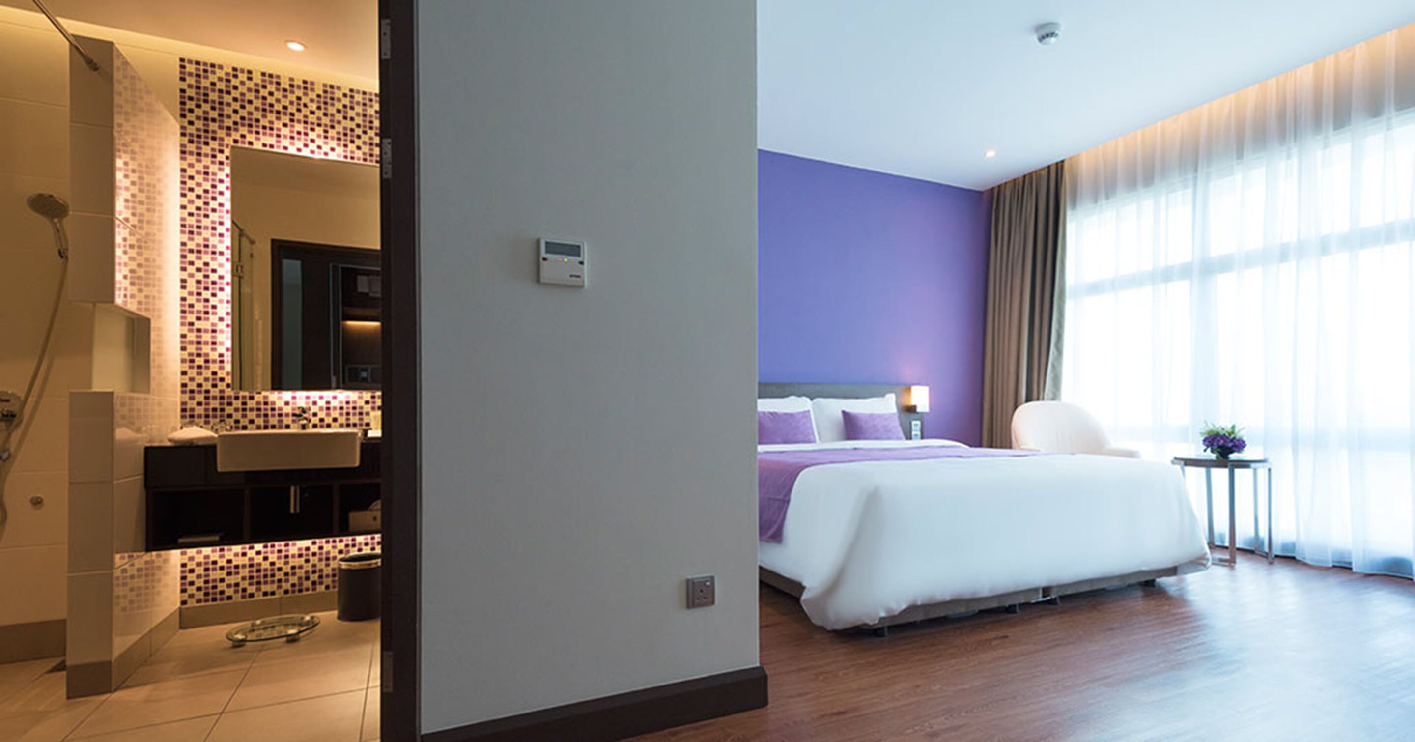 A hotel room with a bathroom, a bed with a purple headboard, a bedside table with a lamp, and a window with curtains.