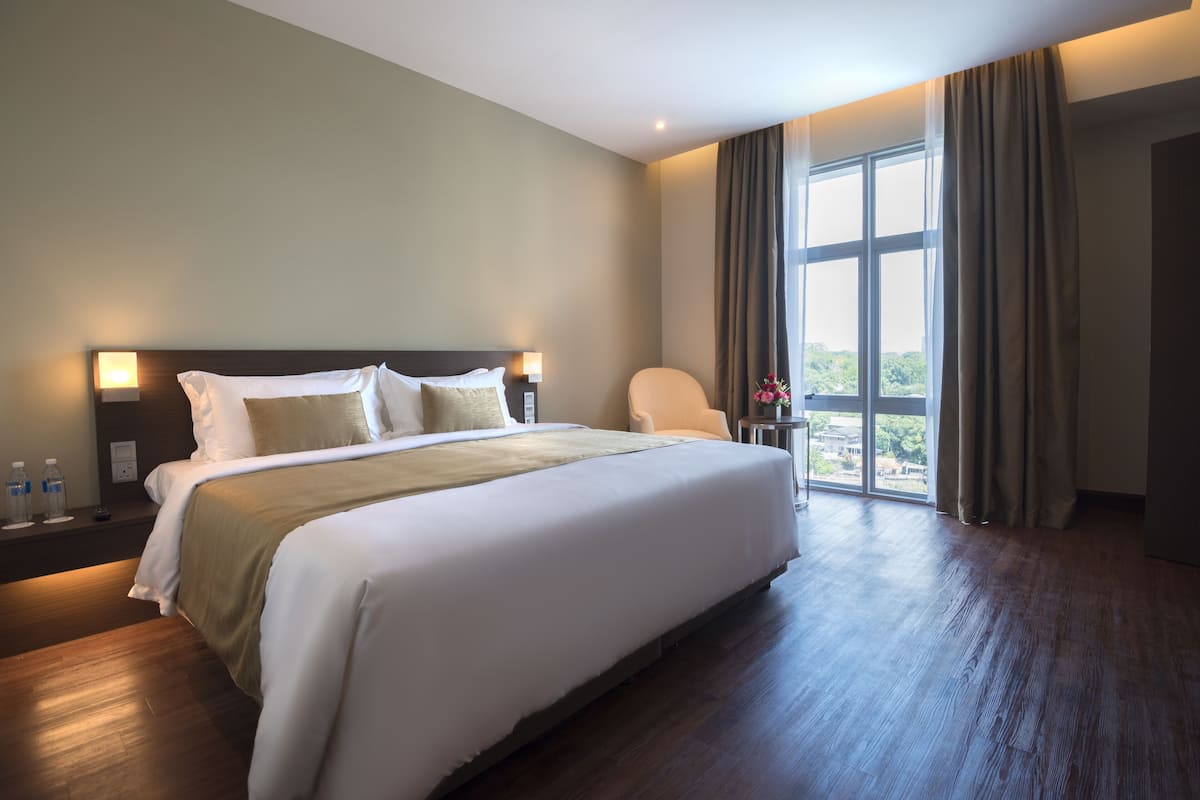 Habitación de hotel con una cama grande, una mesita de noche con lámpara, una silla y vistas al exterior a través de un ventanal amplio.