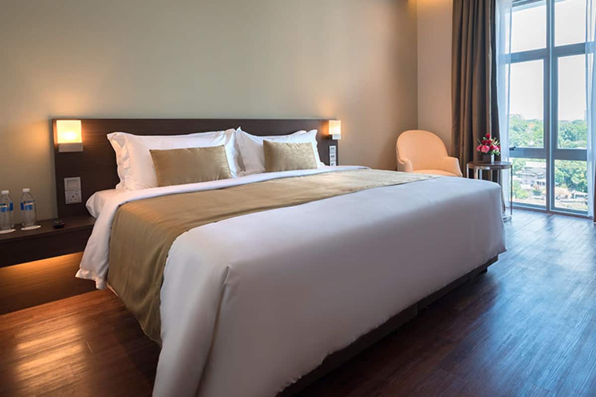 Habitación de hotel con una cama grande, una mesita de noche con botellas de agua, una silla y vistas al exterior a través de un ventanal amplio.