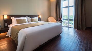 Habitación de hotel con una cama grande, una mesita de noche con botellas de agua, una silla y vistas al exterior a través de un ventanal amplio.