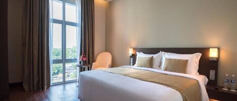 Une chambre d’hôtel avec un grand lit, une chaise, une table de chevet avec une lampe et une vue sur la verdure par la fenêtre.