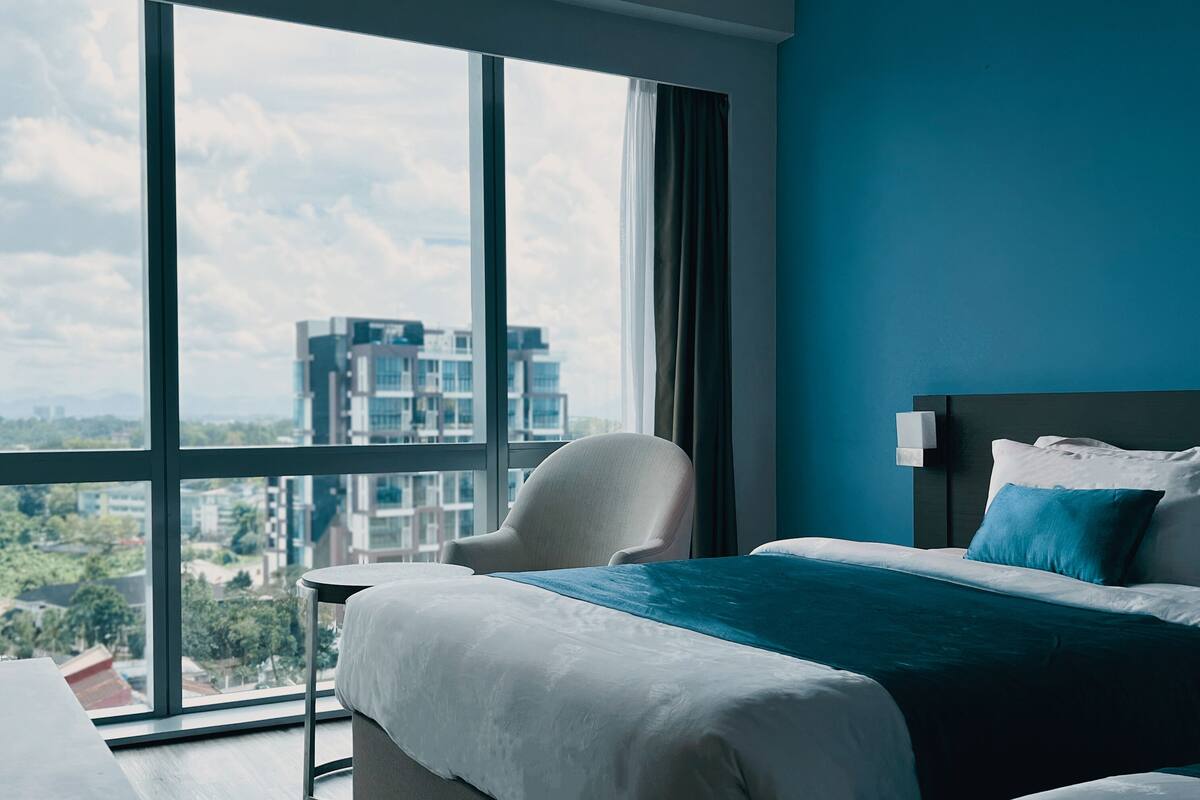 Habitación de hotel moderna con un ventanal grande, cama con sábanas blancas, almohada azul decorativa y vistas a un paisaje urbano.