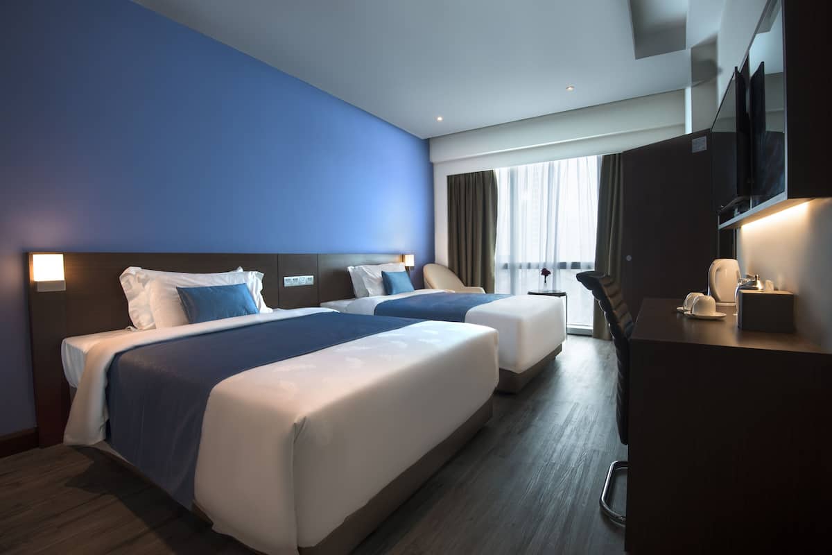 Habitación de hotel con dos camas, un escritorio, un televisor y un ventanal grande con cortinas.