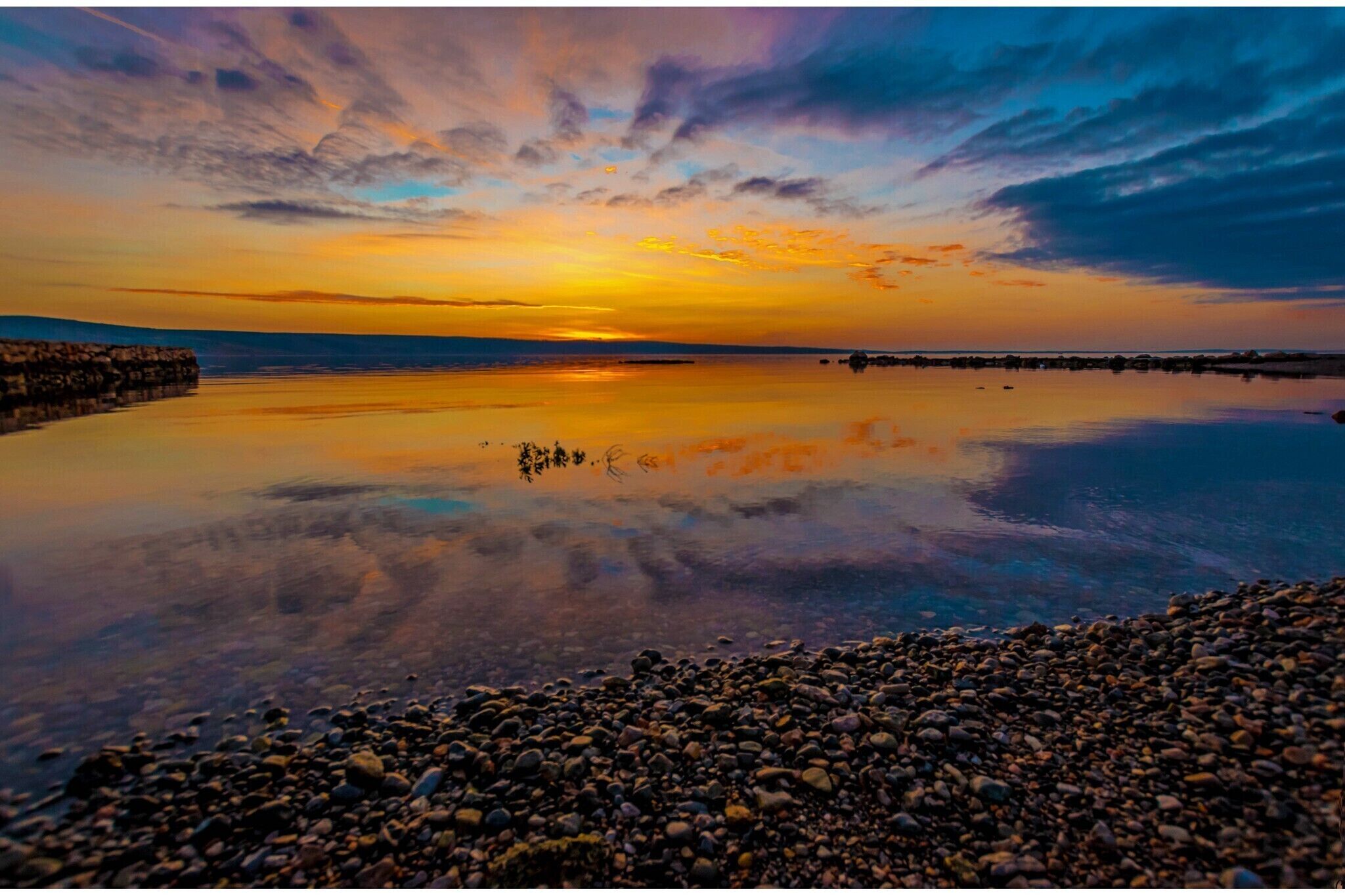 Un coucher de soleil sur une étendue d’eau calme, bordée d’une côte rocheuse.