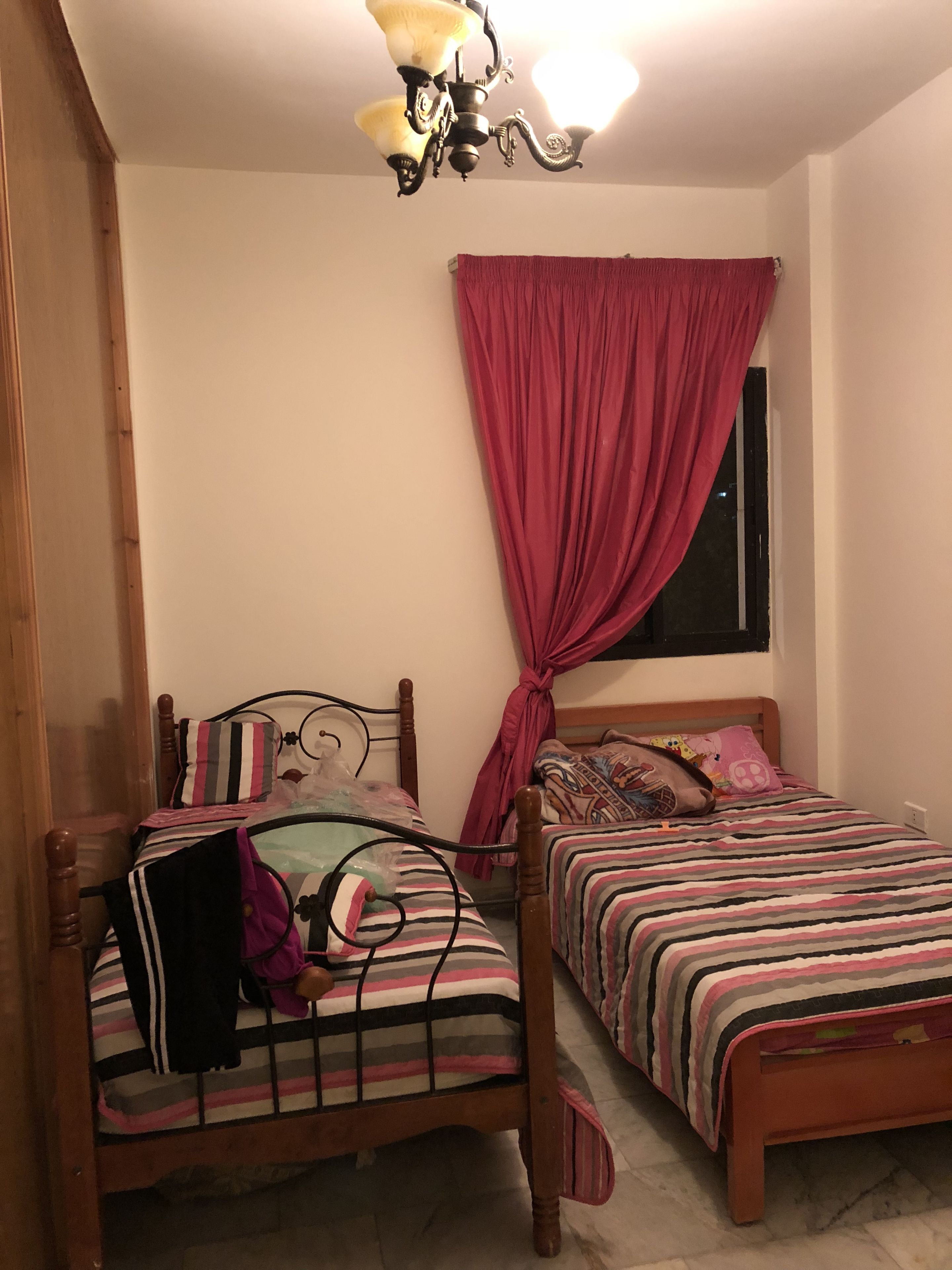 A bedroom with two wooden beds, a window with a red curtain, and a chandelier.