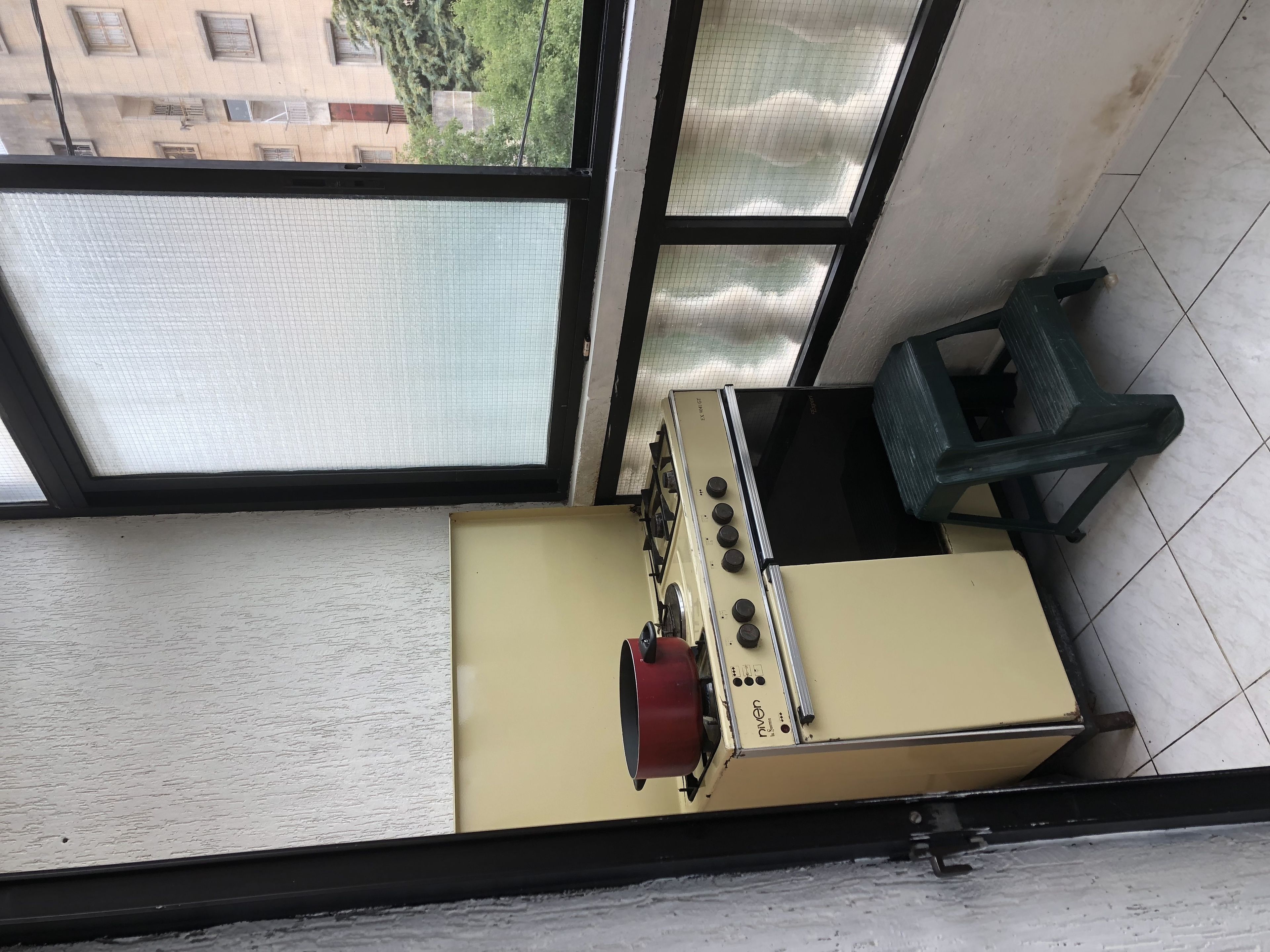 A compact kitchen with a gas stove, a small stool, and a window overlooking a building.