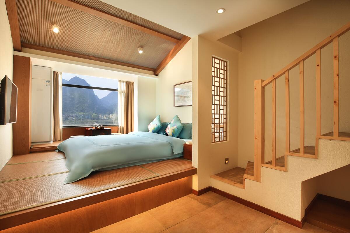 A bedroom with a bed, a window with a view, a wooden staircase, and a tatami mat.