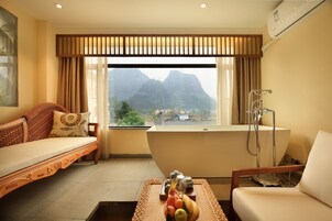 A room with a sofa, a table with fruit, a bathtub, and a view of mountains.