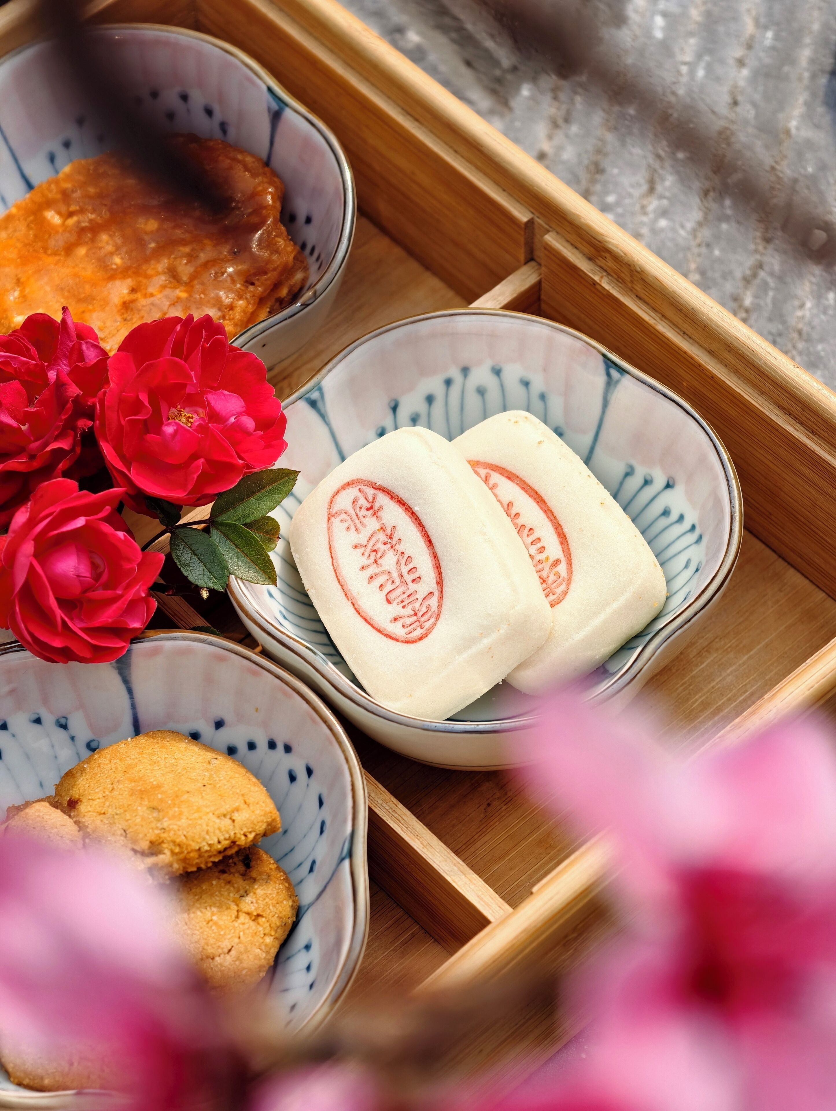 A wooden tray with three bowls containing food items, a red flower, and a pink blurred background.