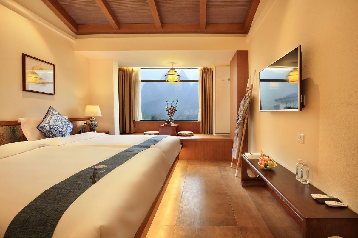 A hotel room with a large bed, a bedside table with a lamp, a small table with a vase of flowers, a mirror on the wall, and a window with curtains.
