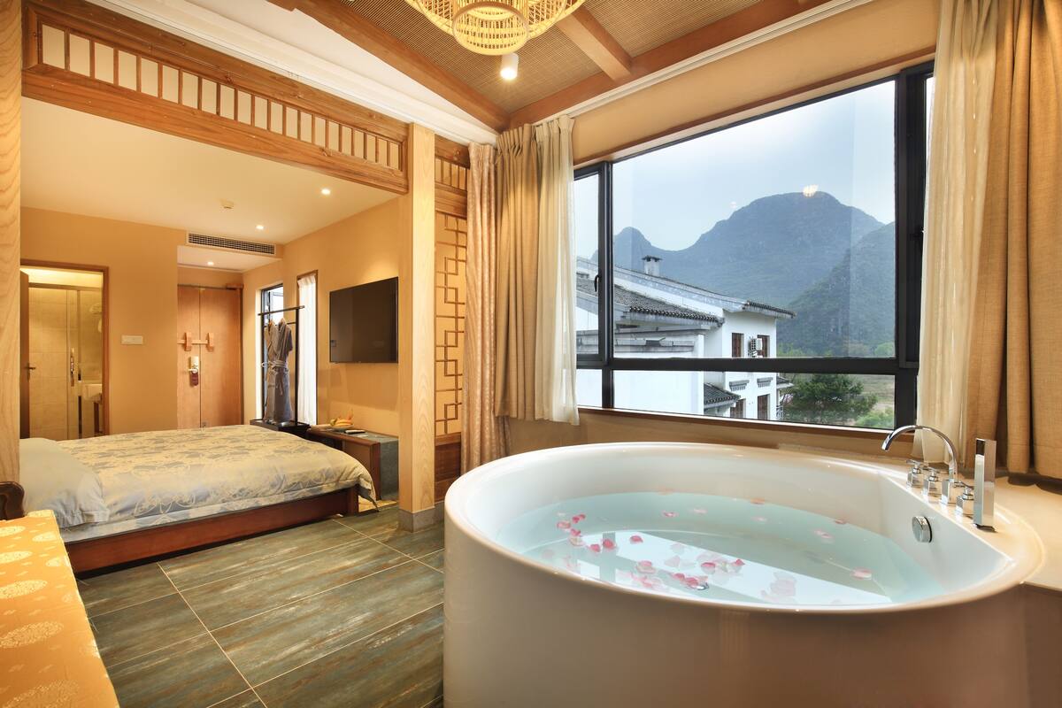 A hotel room with a large bathtub, a bed, and a view of mountains through a window.