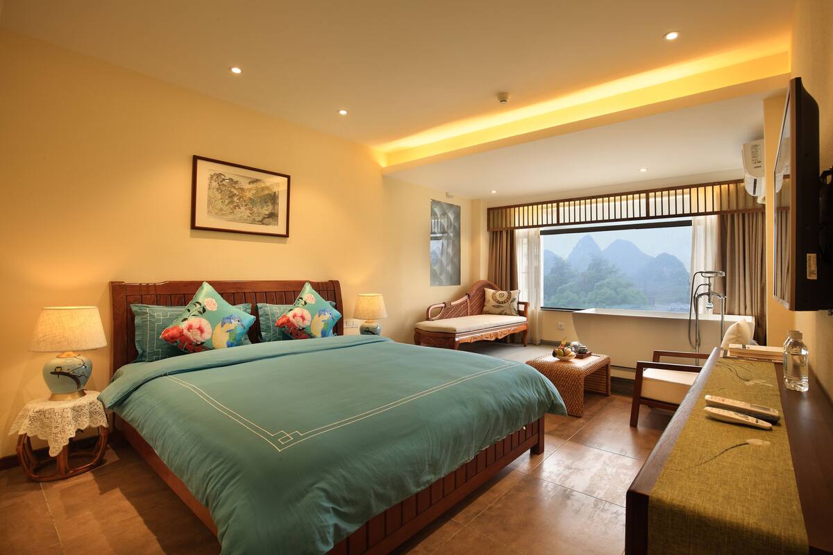 A hotel room with a large bed, a wooden headboard, a bedside table with a lamp, a small table with fruit, a bench, and a view of mountains through the window.