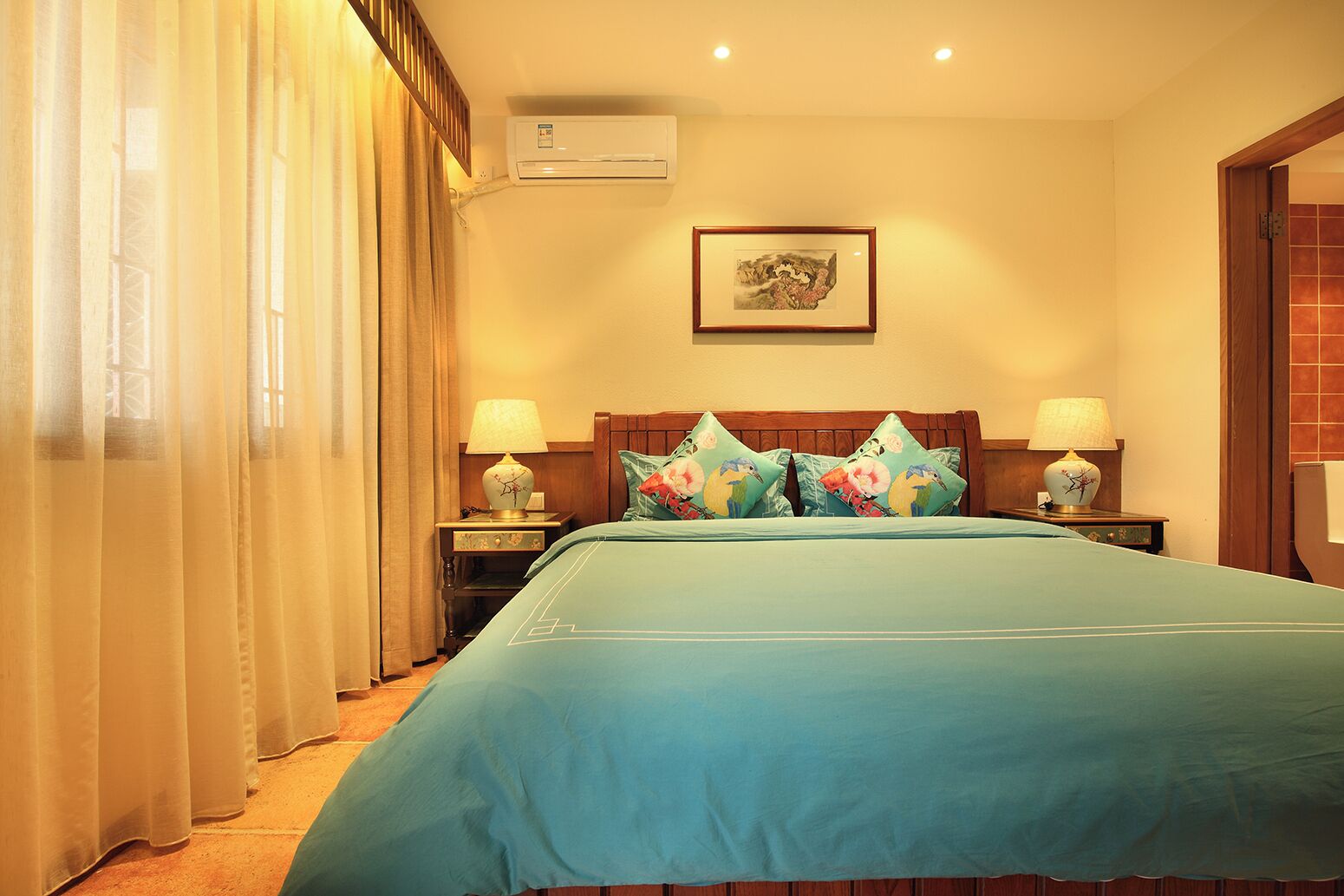 A hotel room with a bed, two bedside tables with lamps, a wall-mounted air conditioner, and a framed picture above the bed.