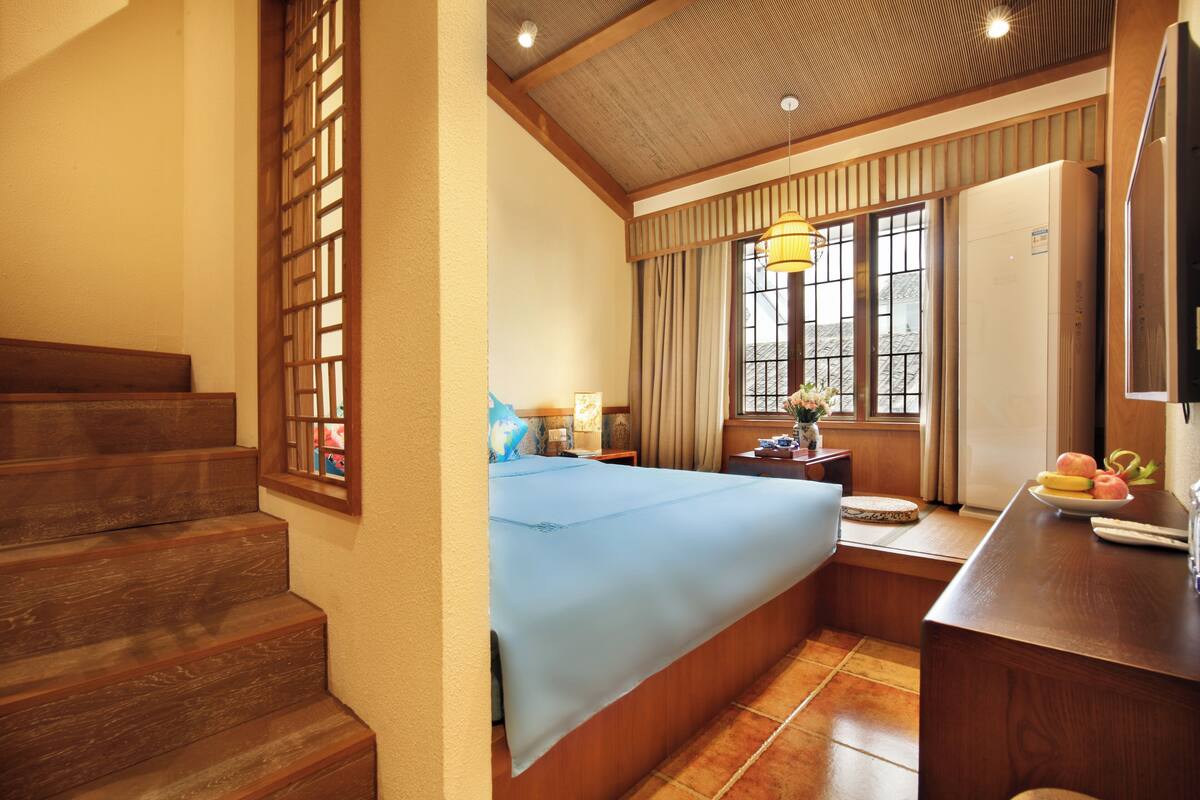 A hotel room with a wooden staircase, a bed with a blue bedspread, a desk with a fruit bowl, and a large window with curtains.