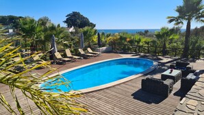 Ein Poolbereich mit Liegestühlen und Sonnenschirmen, umgeben von Palmen und Blick aufs Meer.