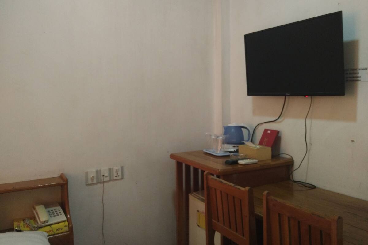 A small room with a wooden desk, a chair, a bed, a wall-mounted TV, a telephone, and a power outlet.