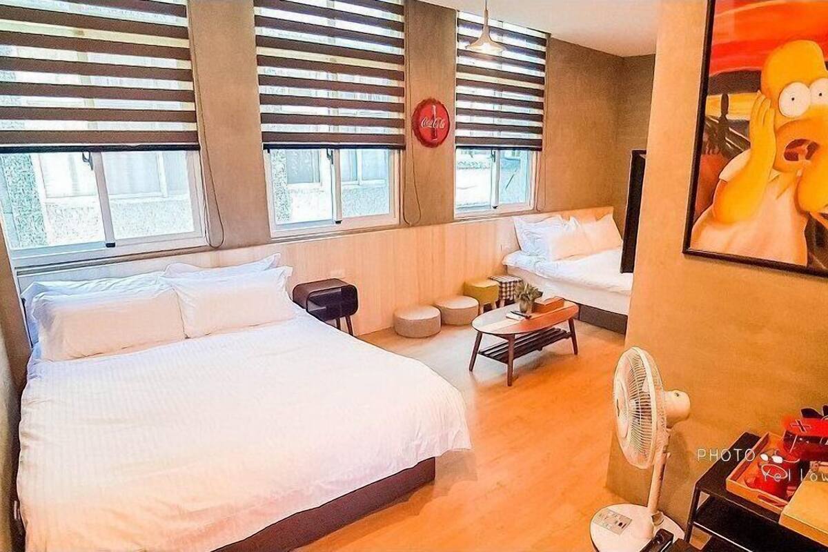 A hotel room with two beds, a sofa, a small table, a fan, and a painting on the wall.