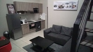 A compact living space with a kitchenette, a grey sofa, a coffee table, and a staircase. - Apartment In Rosebank Johannesburg (Johannesburg)
