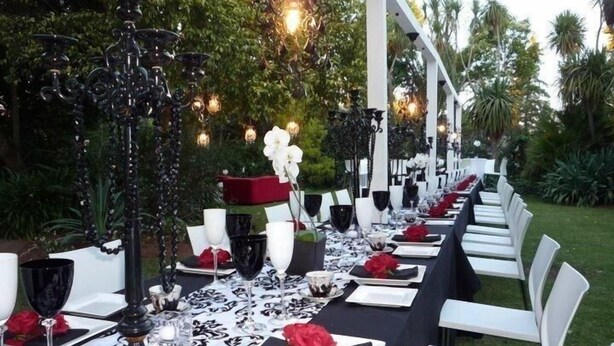 Un área de comedor al aire libre con una larga mesa dispuesta para un evento formal, rodeada de vegetación e iluminación decorativa.