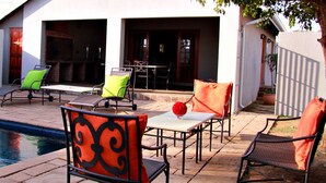 Zona de piscina con una pileta rectangular, mobiliario exterior con sillas y mesas, y entrada de edificio con puerta y ventanas.