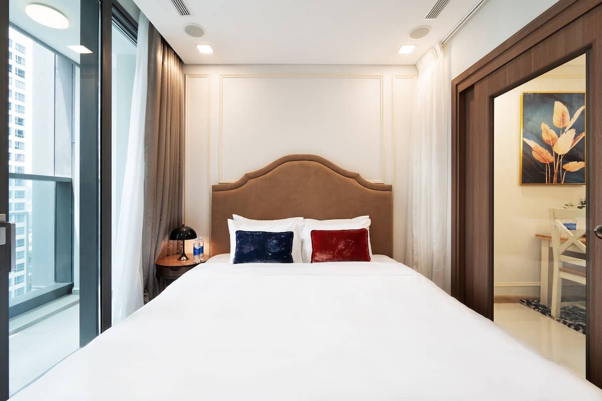A hotel room with a large bed, a nightstand, a lamp, and a view of the city.