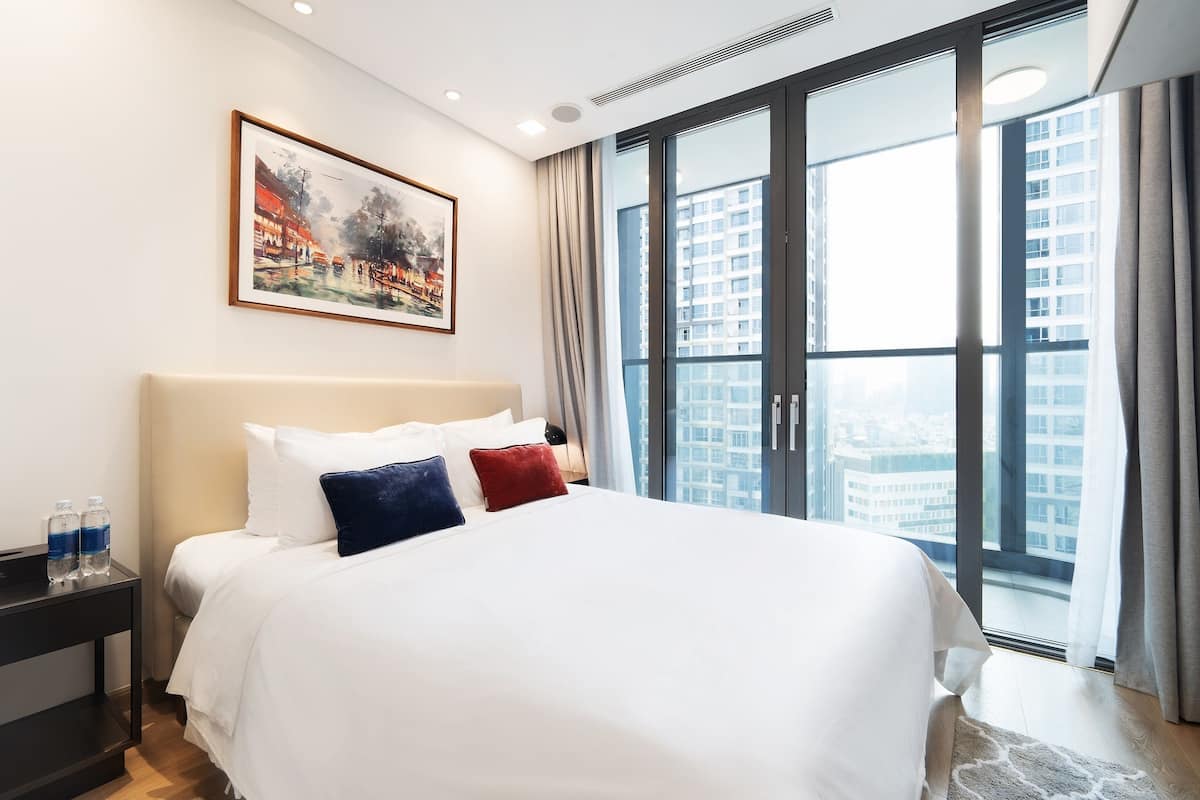 A hotel room with a large bed, a painting on the wall, and a view of a cityscape through a large window.