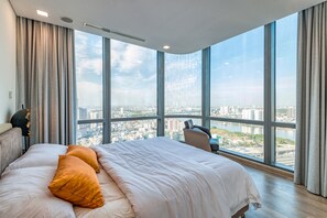 Une chambre d’hôtel moderne dotée d’un grand lit, offrant une vue sur le paysage urbain grâce à de larges fenêtres du sol au plafond, et permettant d’apercevoir la silhouette de la ville au loin.