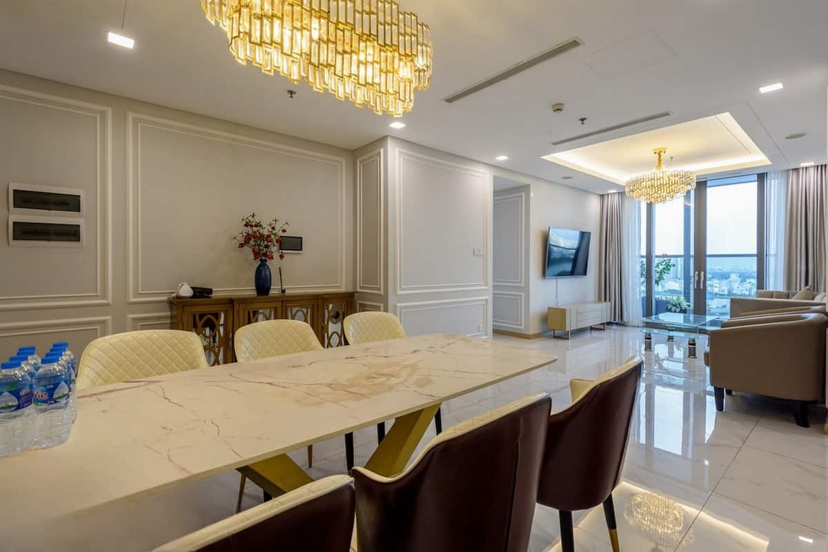 A modern hotel room with a large marble dining table, comfortable seating, a chandelier, a flat-screen TV, and a city view.