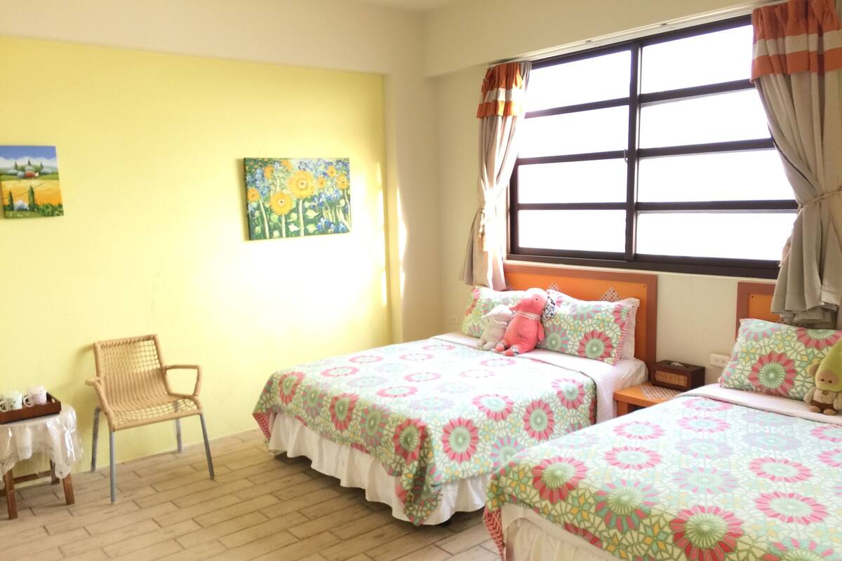 Quarto com duas camas, uma cadeira, uma mesa pequena e uma janela com cortinas.