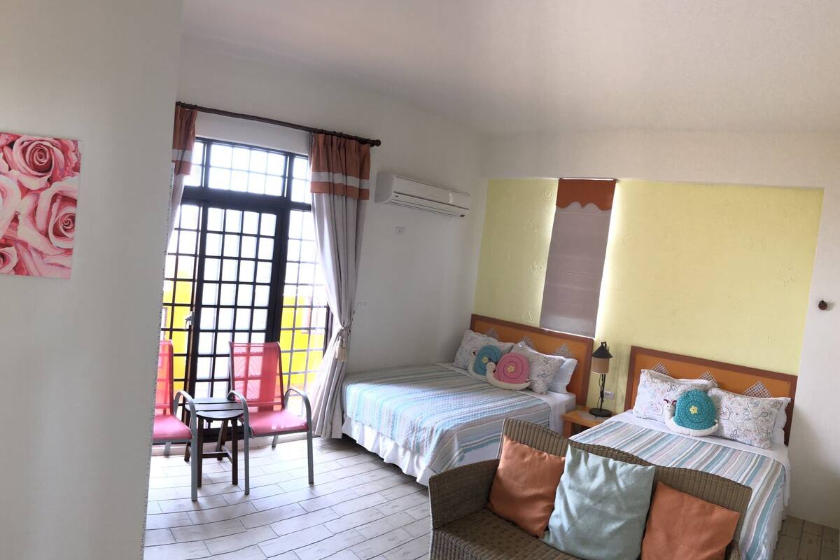 Quarto com duas camas, janela com cortinas, um ventilador e um quadro de rosas.